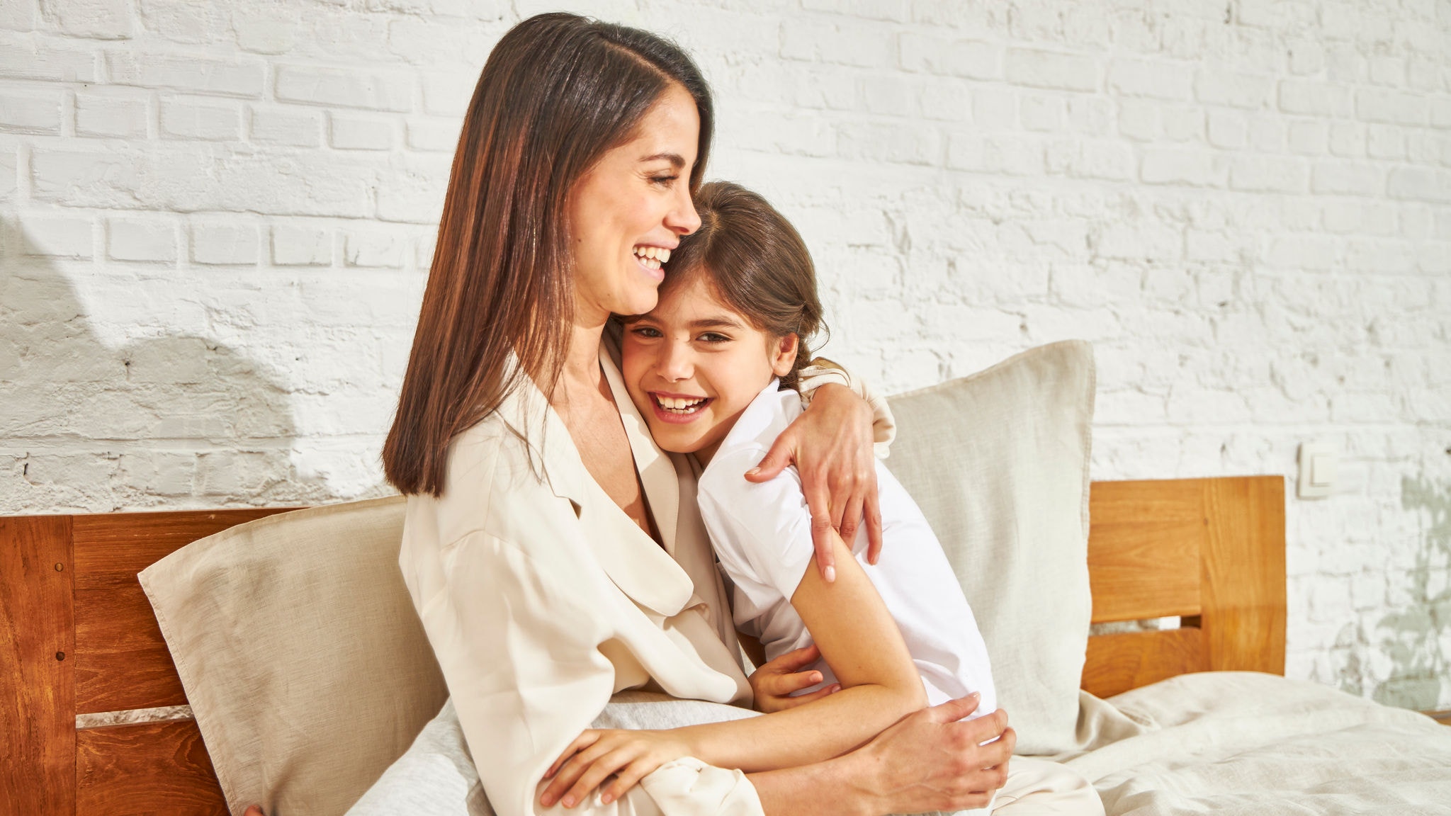 Mother and little daughter smiling and hugging while sitting on a bed