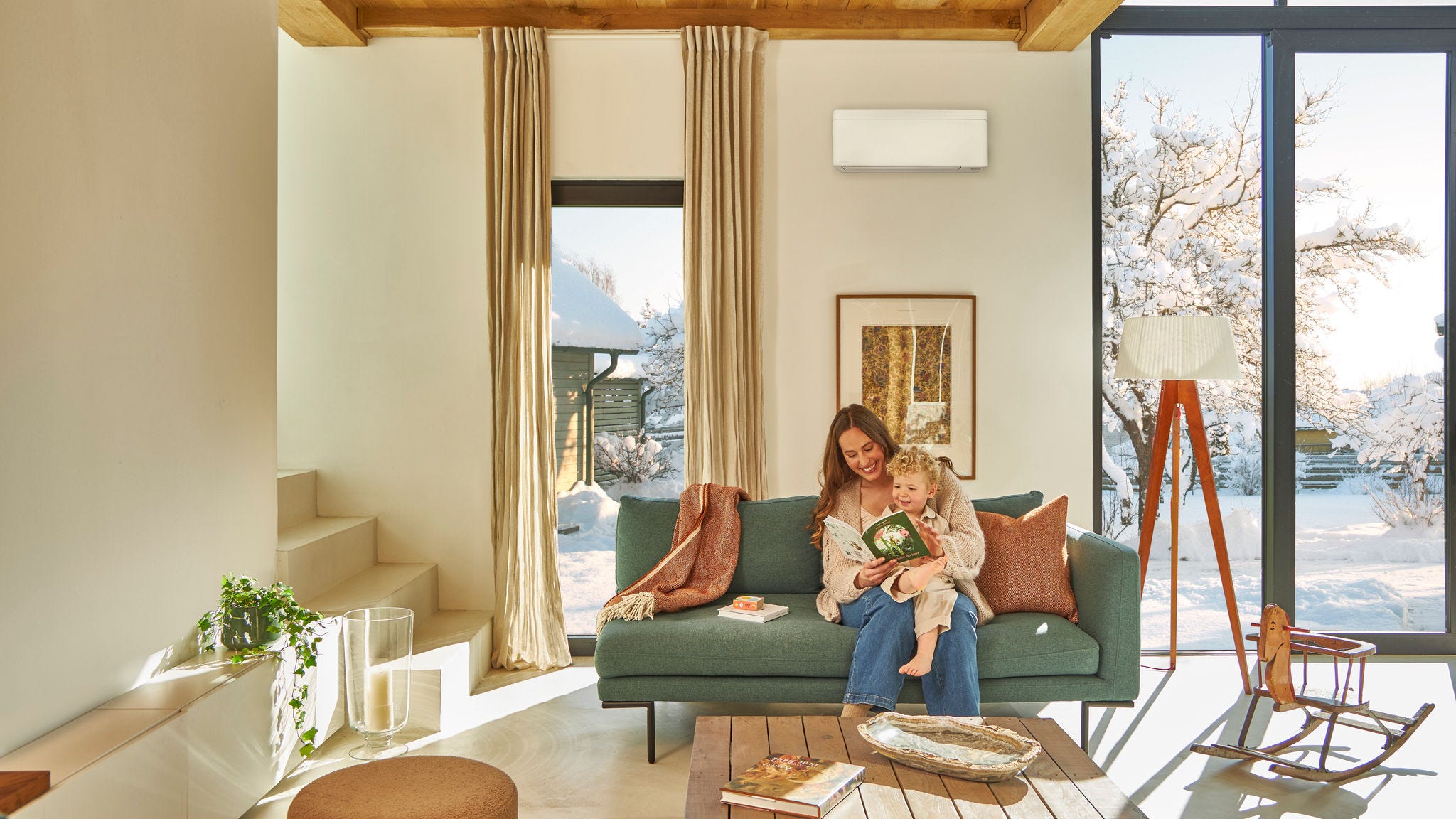Mère assise sur le canapé sous une pompe à chaleur air/air murale, lisant un livre à son enfant assis sur ses genoux, profitant de la chaleur alors qu'il neige dehors.